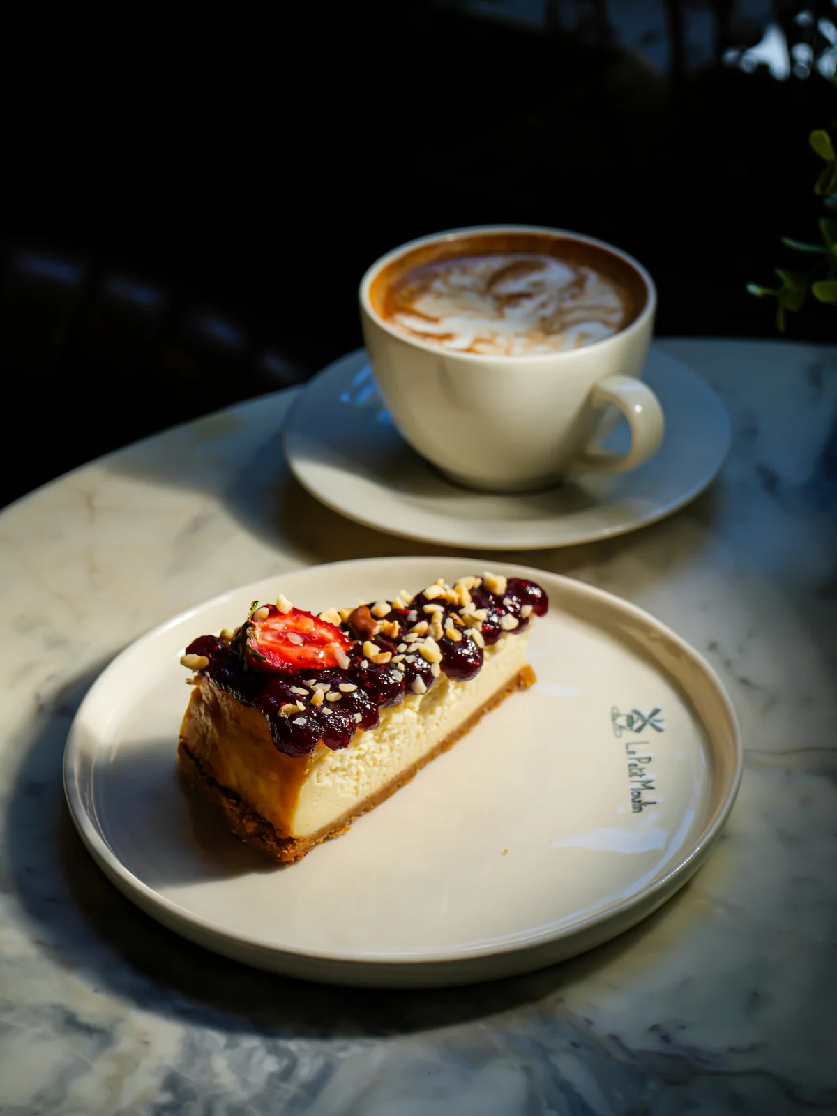 Le Petit Moulin'in Bakırköy Zeytinlik'teki mermer masasında logolu tabakta cheesecake dilimi: üzerinde kırmızı meyve sosu, fındık kırıkları ve çilek; arkada cappuccino